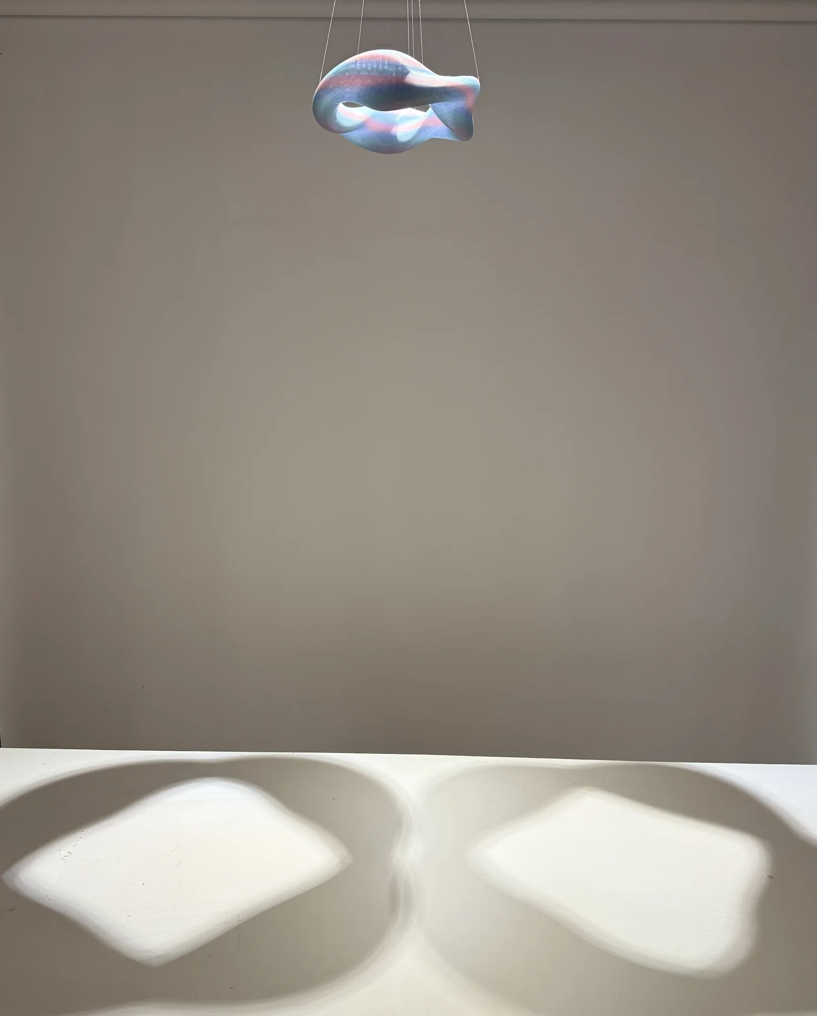 A small suspended form casts two large shadows on a white plinth.