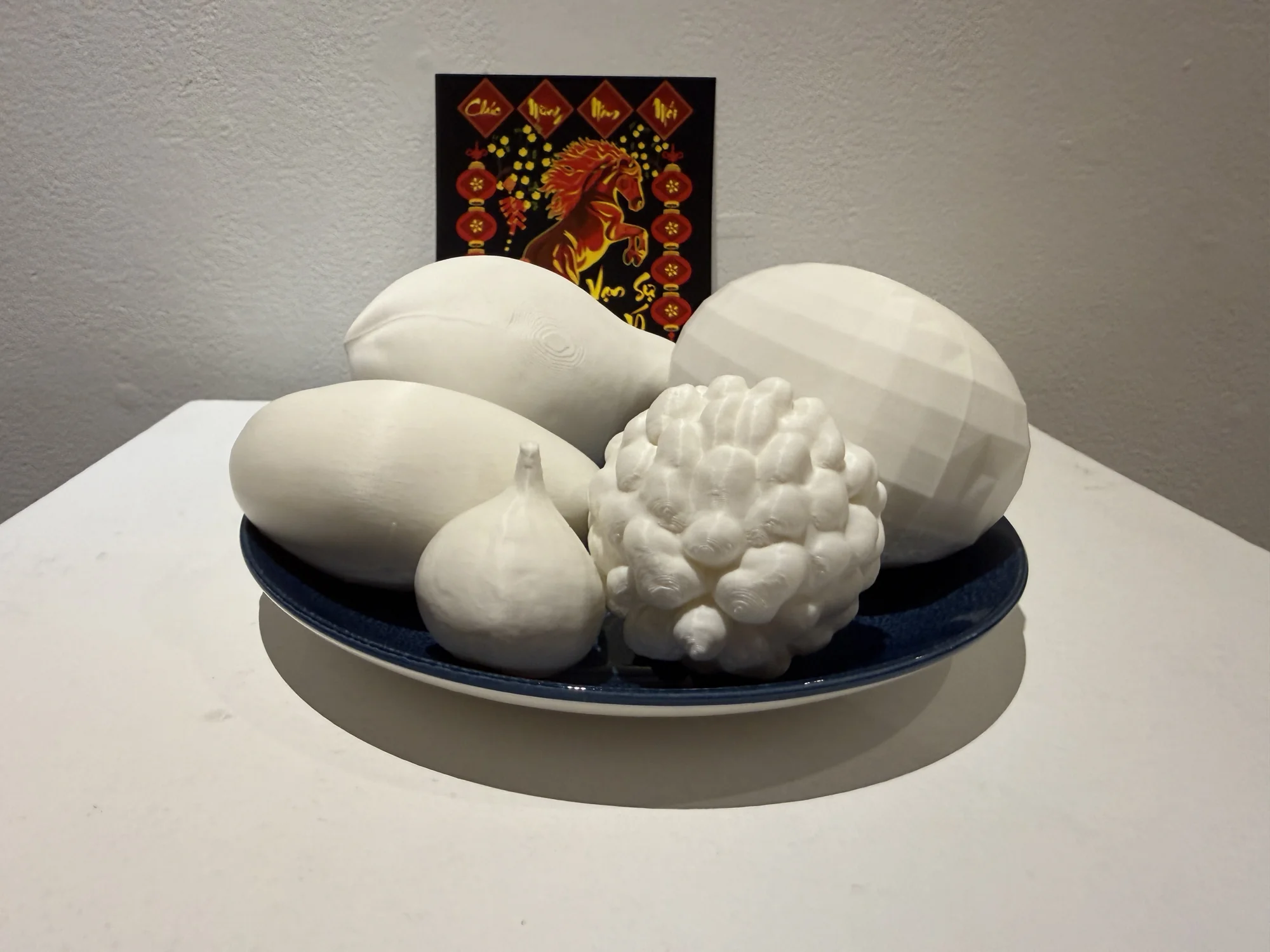 White fruit and vegetable forms arranged on a blue plate beneath a small wall image.
