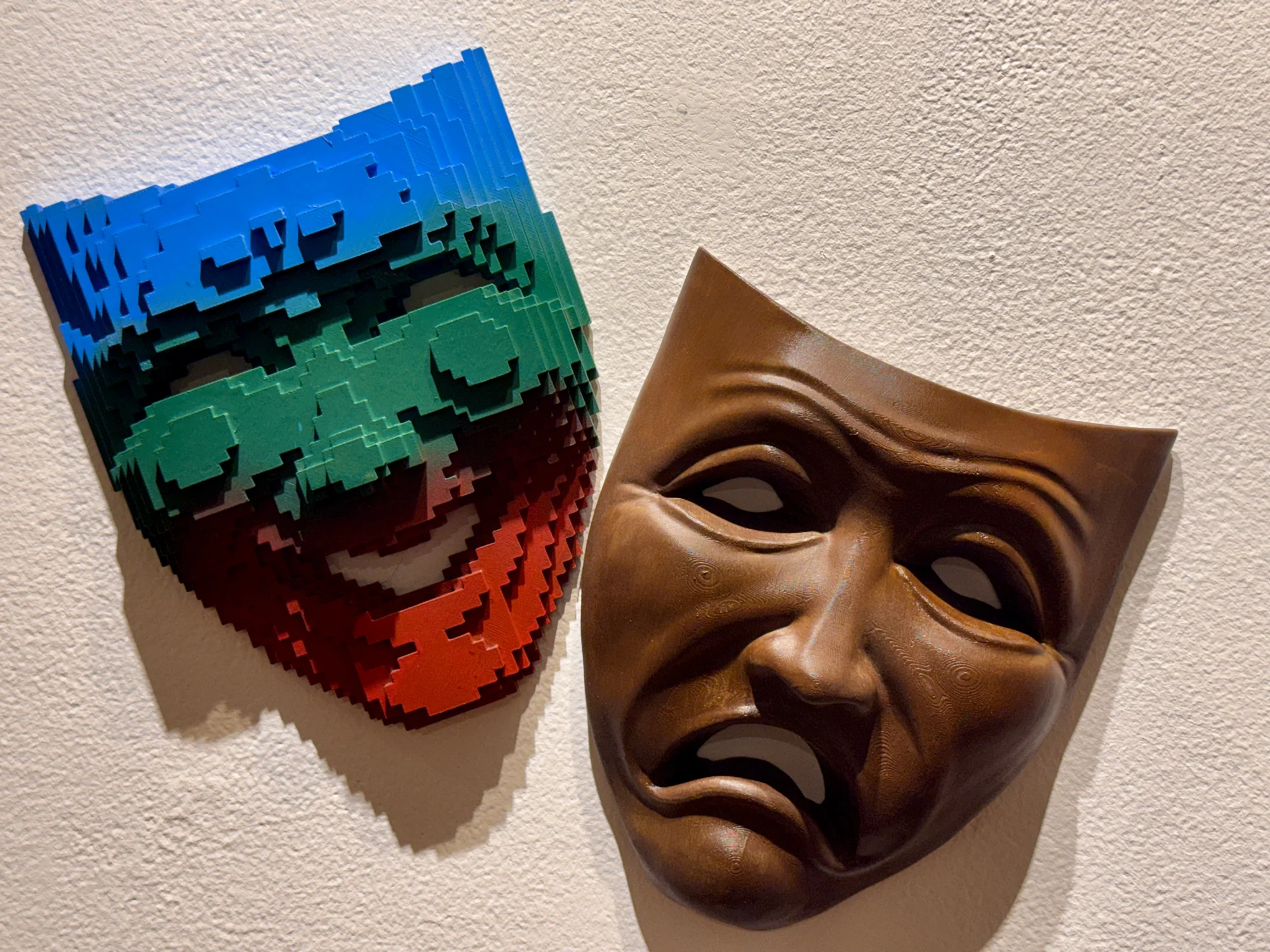 A colorful pixelated mask displayed beside a bronze-colored theatrical mask.