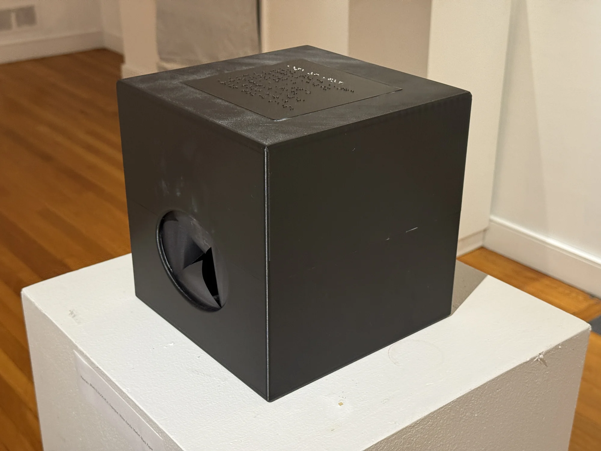 A black cube with a circular opening rests on a white plinth.